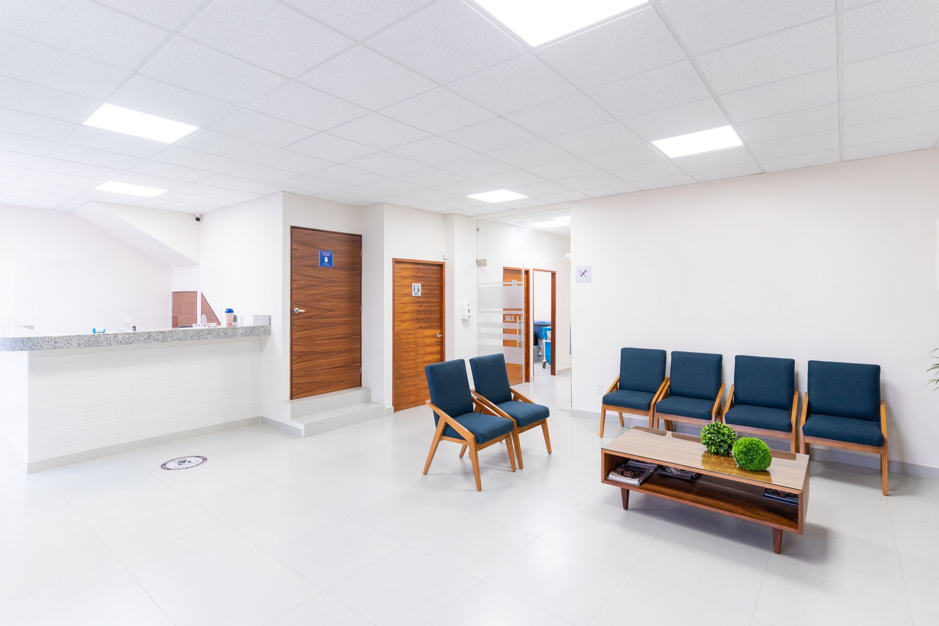 A bright, clean medical waiting room with blue chairs, a coffee table, and a reception counter