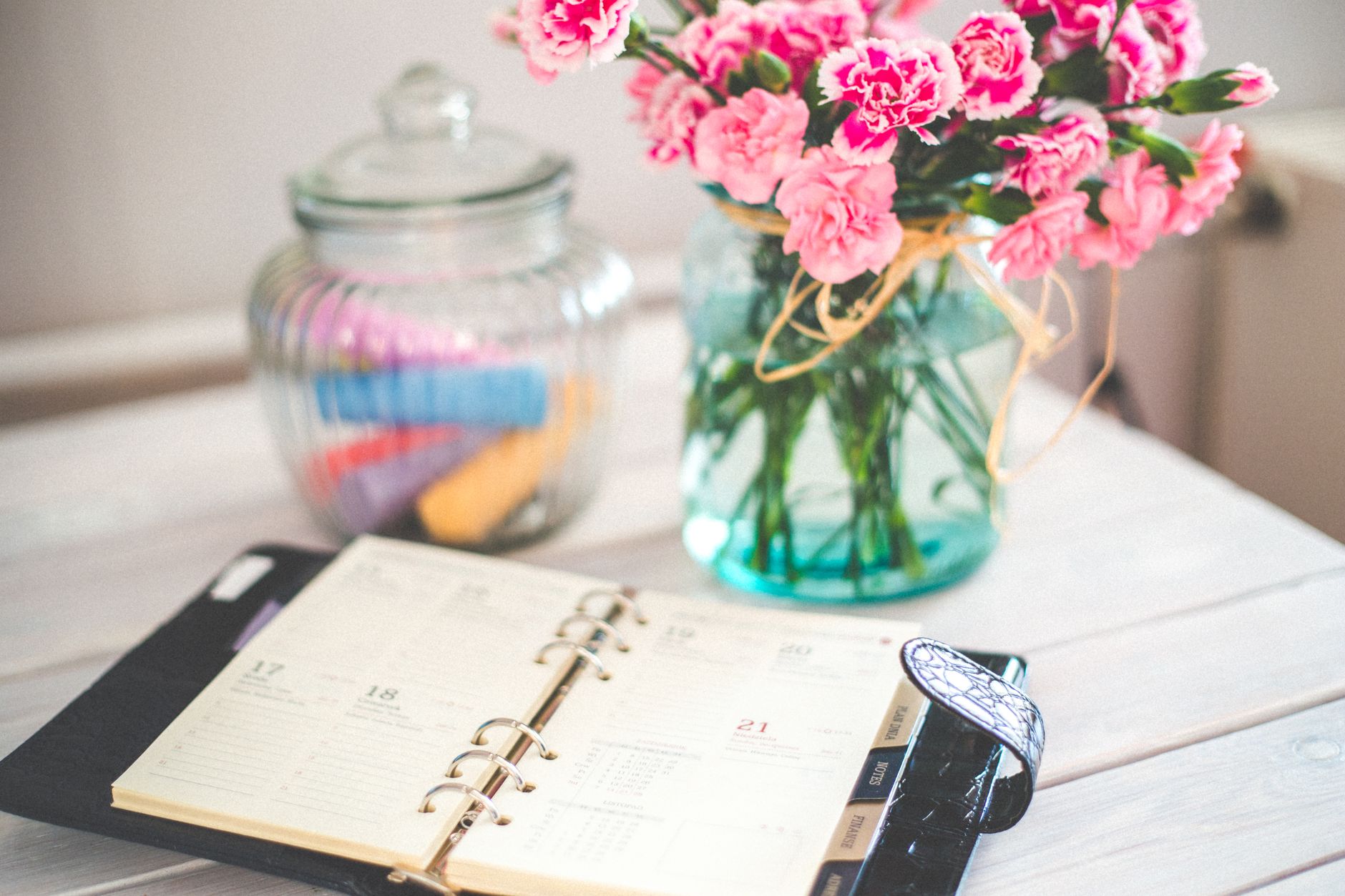 An open planner beside a jar of pink carnations in a glass vase on a white desk