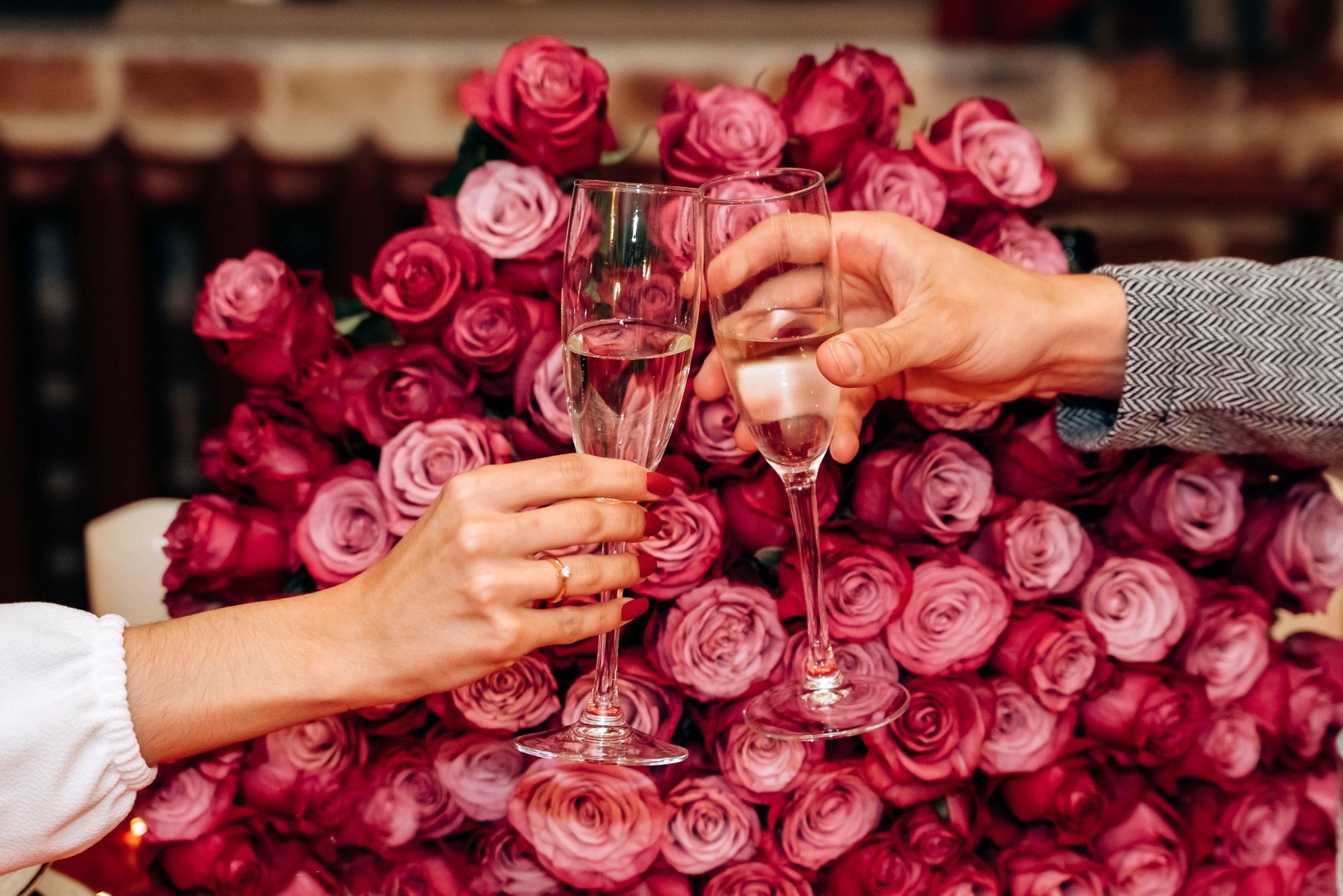 Two hands toasting with champagne flutes in front of a large mass of pink roses