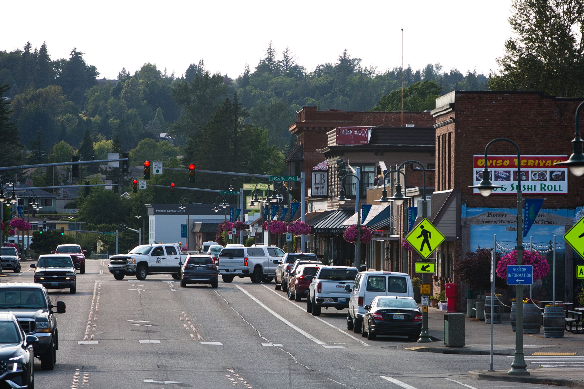 A small-town main street with brick shops, hanging flower baskets, parked cars, and forested hills behind