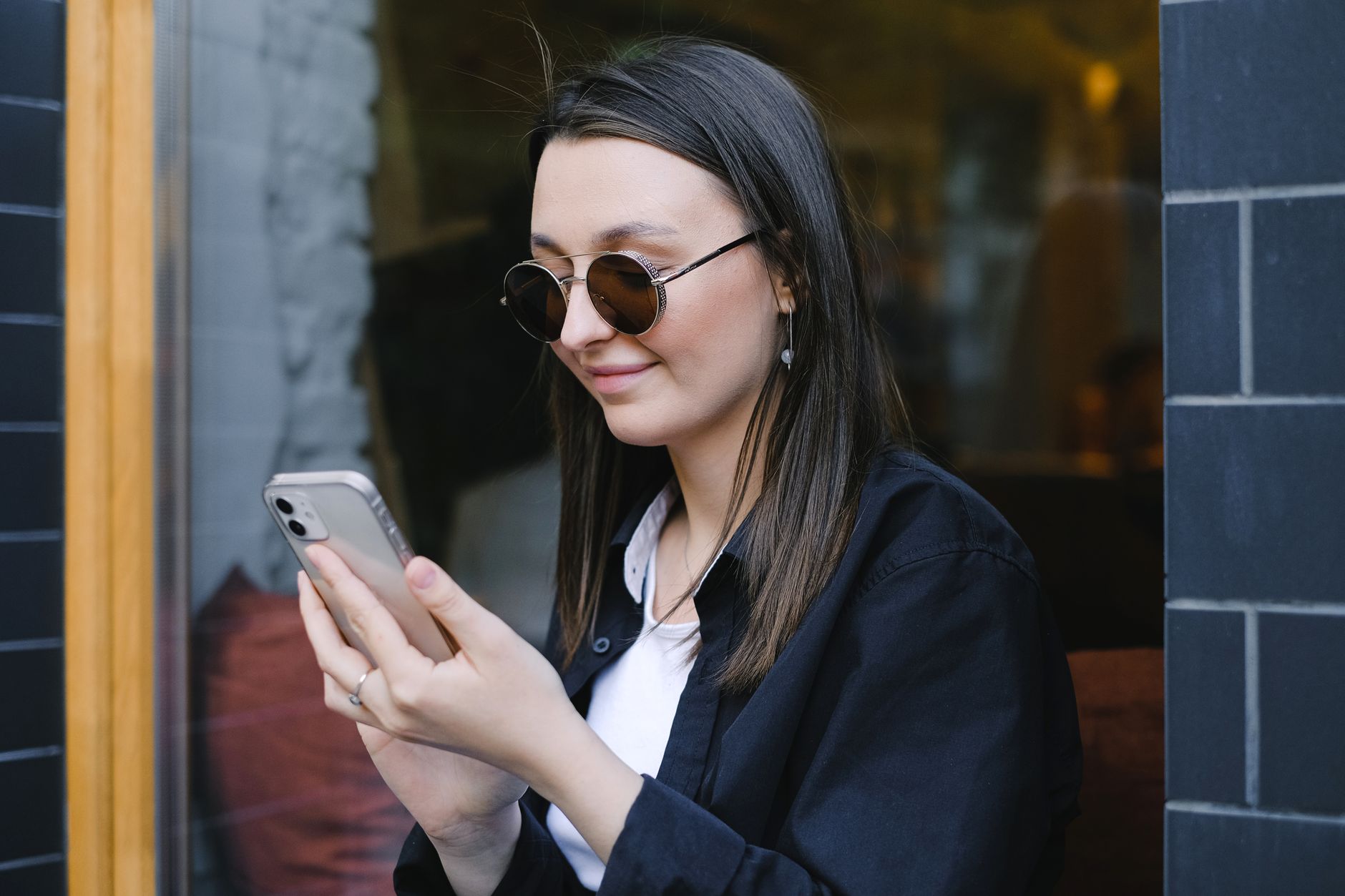 A woman in round sunglasses smiling at her phone outside a cafe window
