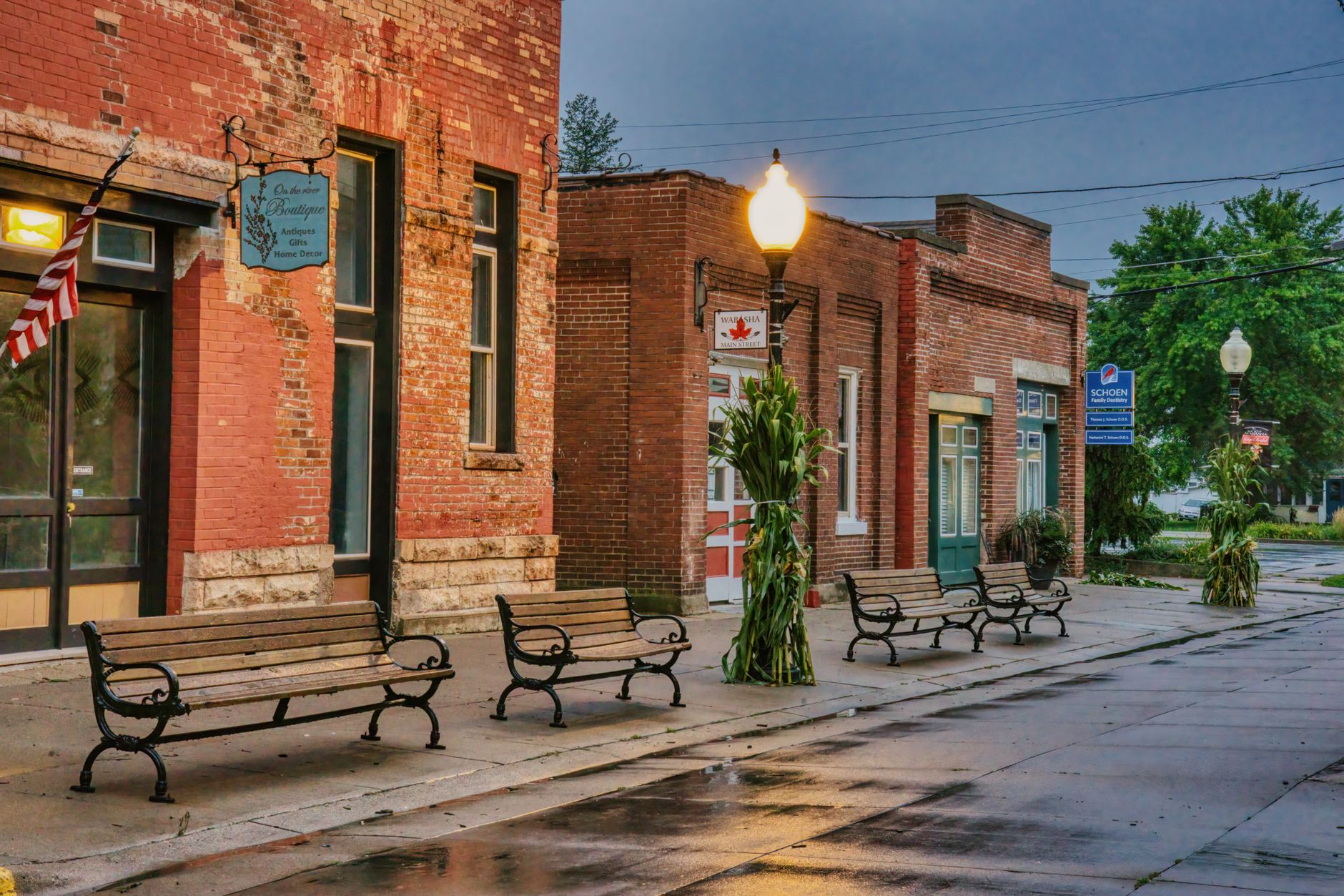 A historic brick main street with benches, corn stalks, a flag, and old storefronts after rain