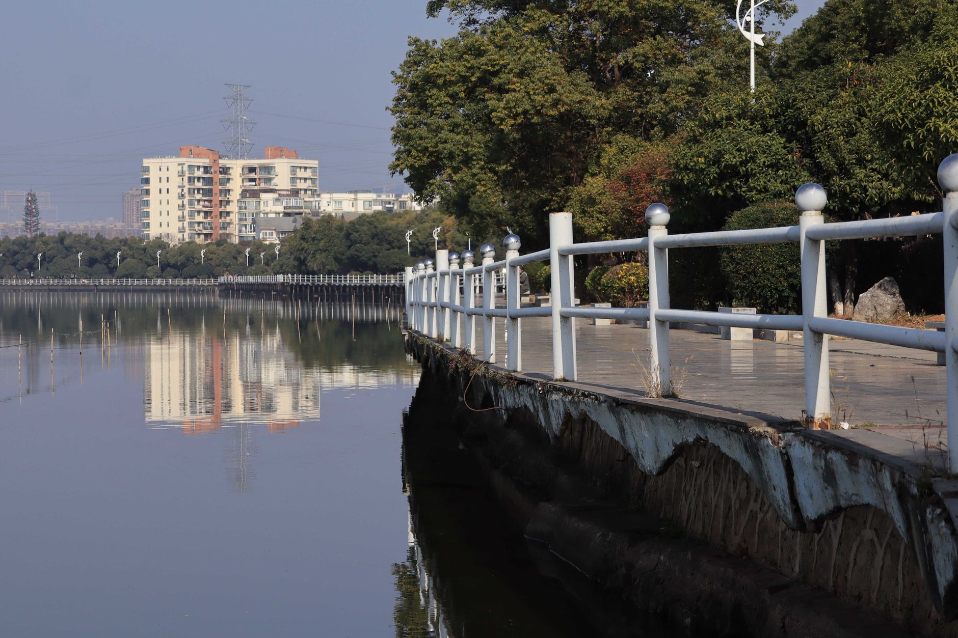 A white railing walkway beside a calm lake reflecting trees and apartment buildings