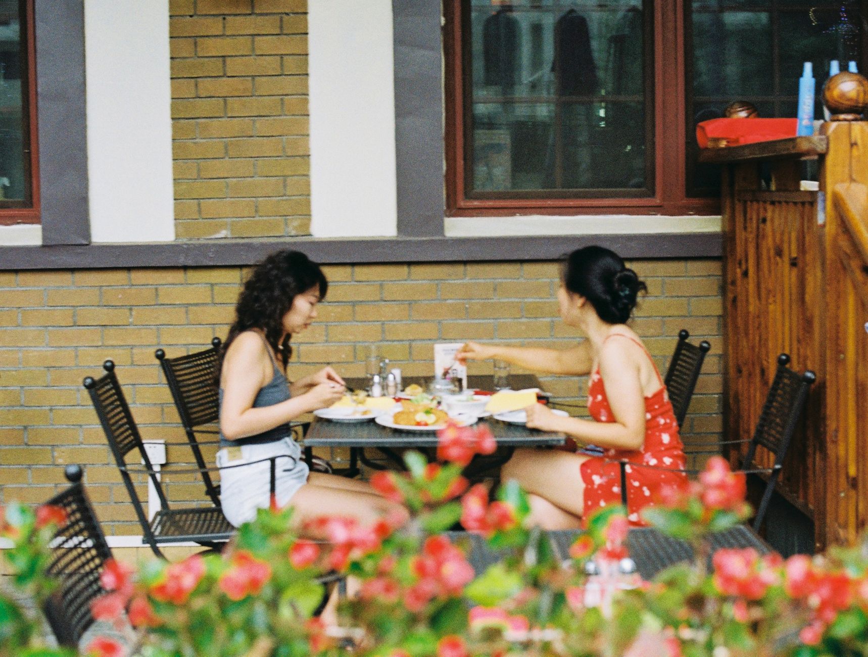 Two women eating at an outdoor patio table behind a hedge of red flowers