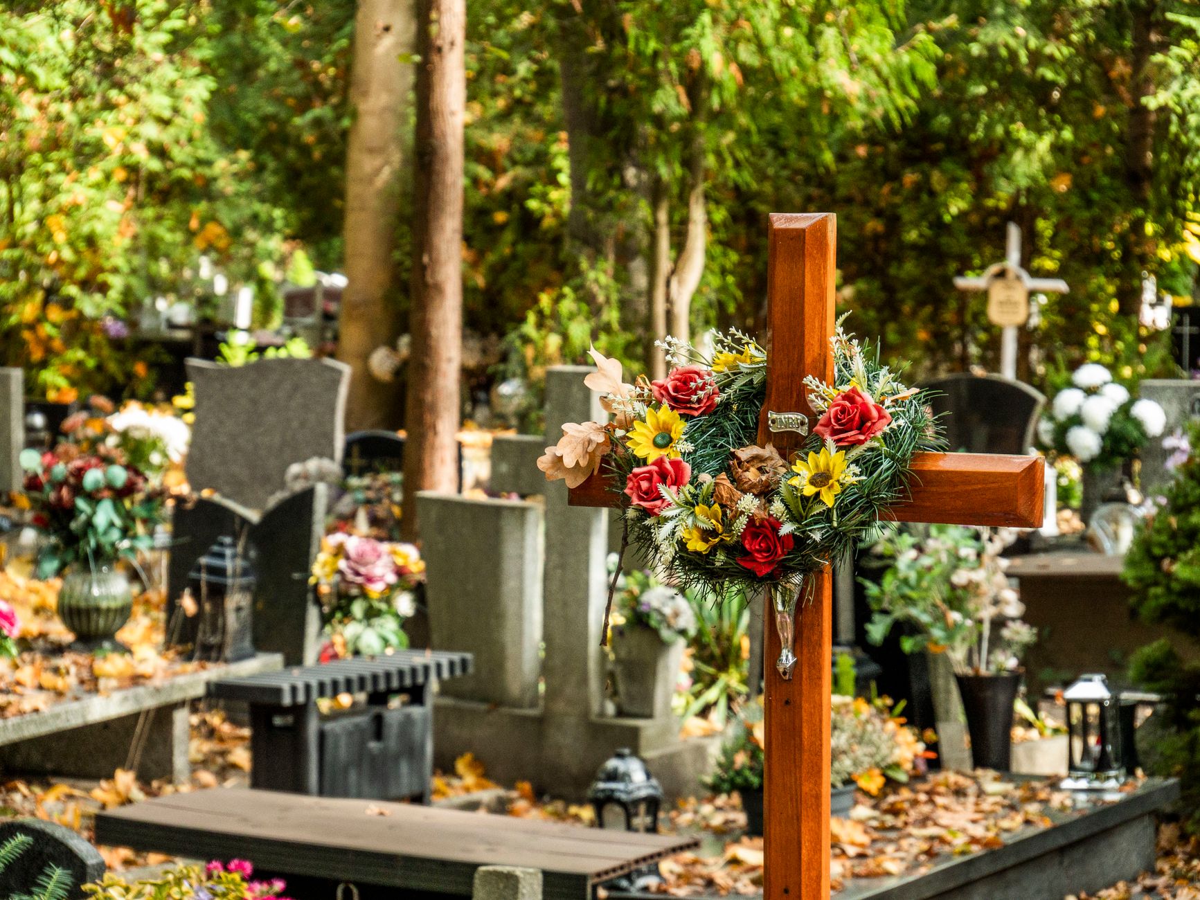 A wooden cross decorated with yellow and red flowers in a cemetery with autumn leaves