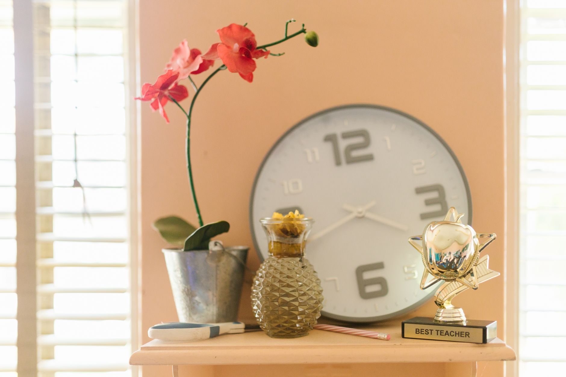 A shelf with a pink orchid, a clock, a glass vase, and a Best Teacher trophy