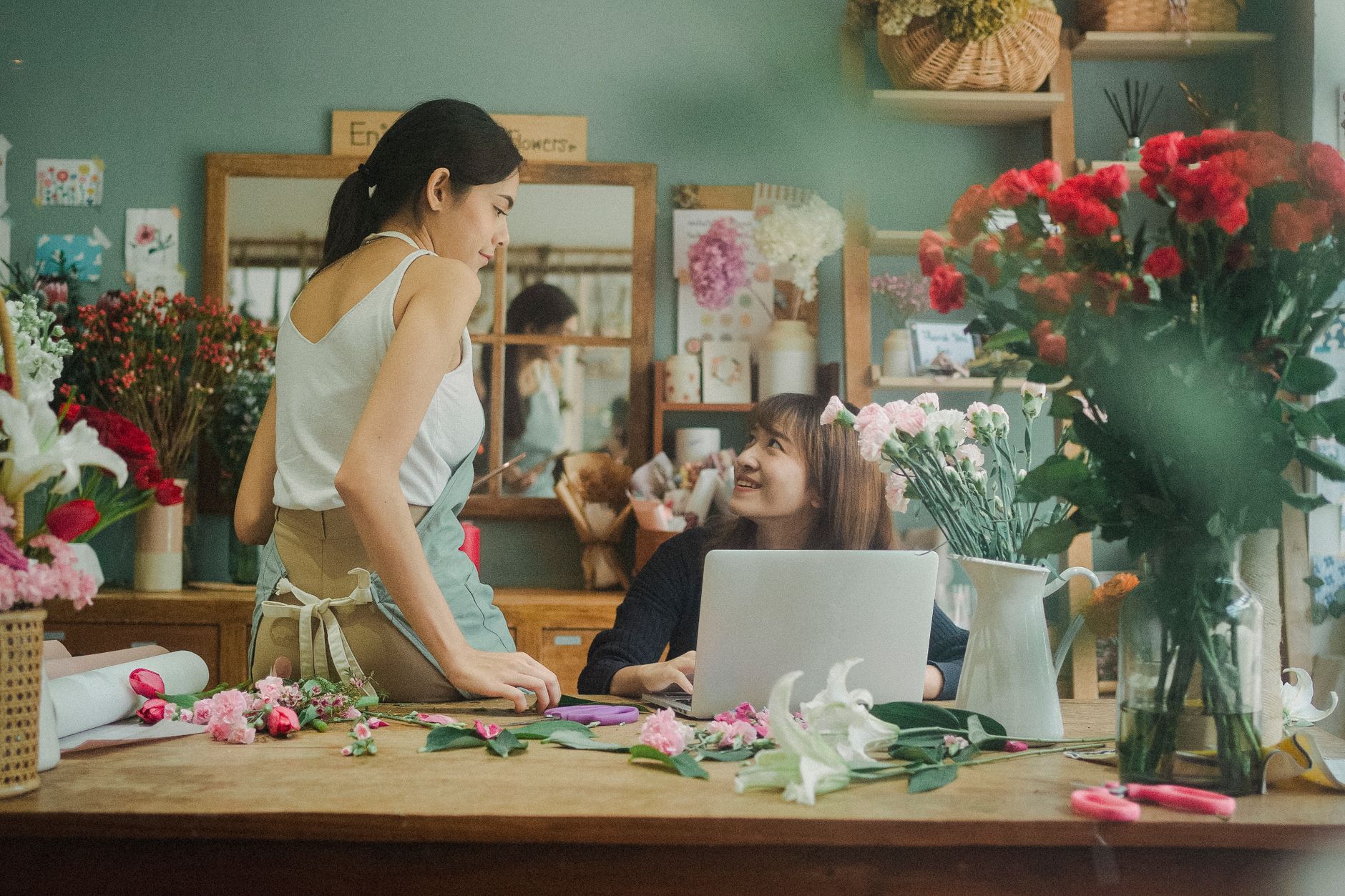 Two women at a flower shop table with bouquets in front of them, looking at a tablet