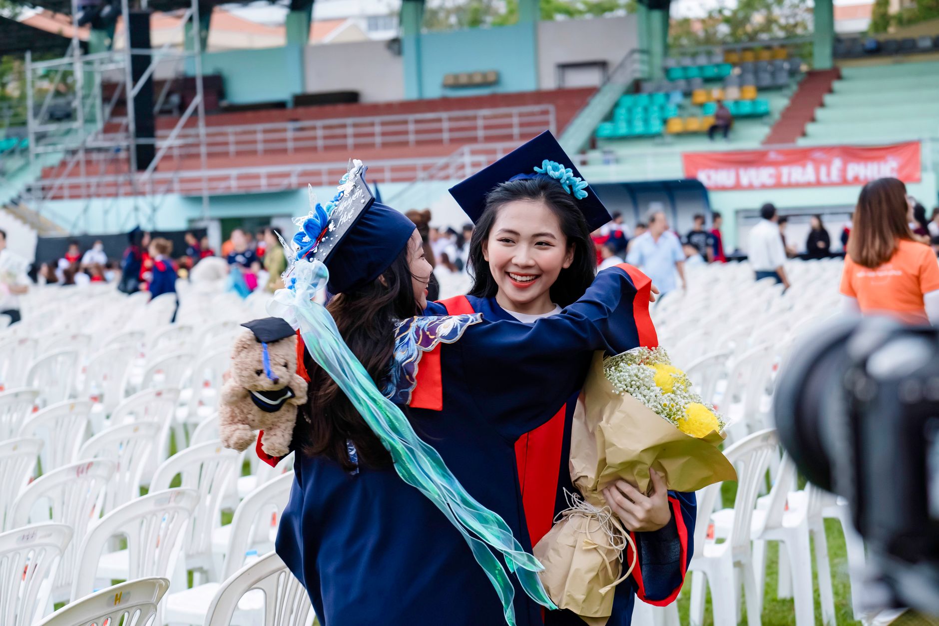 Two graduates in blue gowns hugging at an outdoor ceremony with a bouquet and a stuffed bear