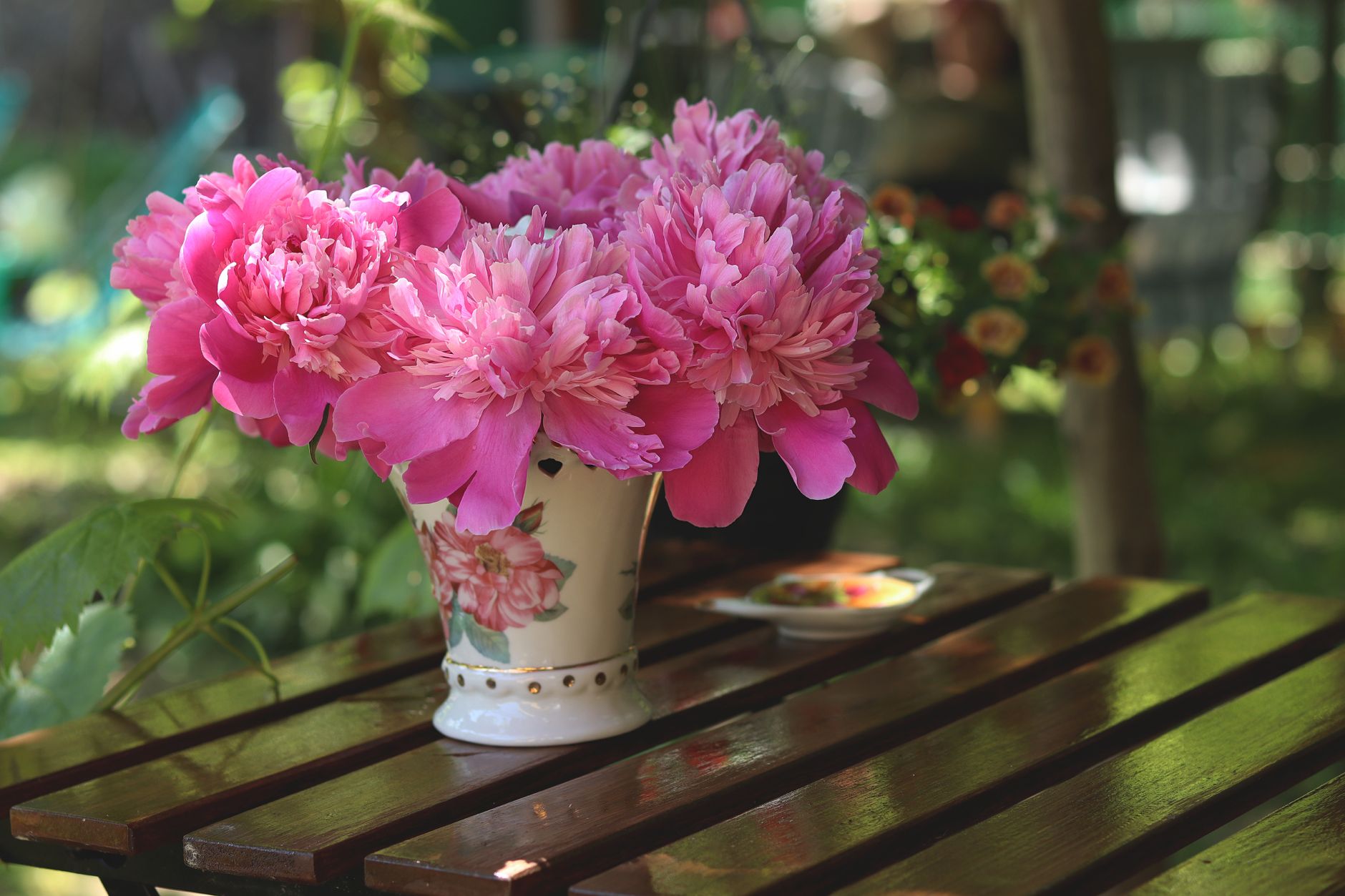 Pink peonies in a flowered ceramic pitcher on a wooden outdoor table in the sun