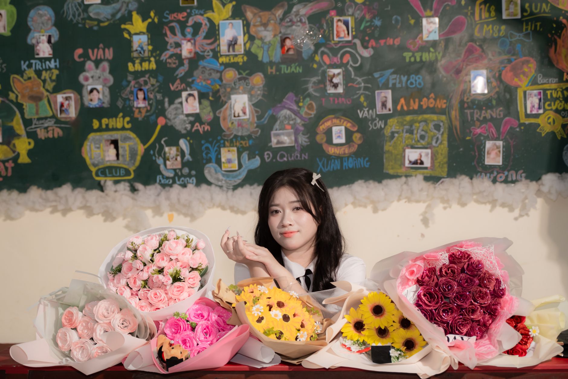 A student at a classroom desk surrounded by bouquets of pink roses, sunflowers, and mixed flowers
