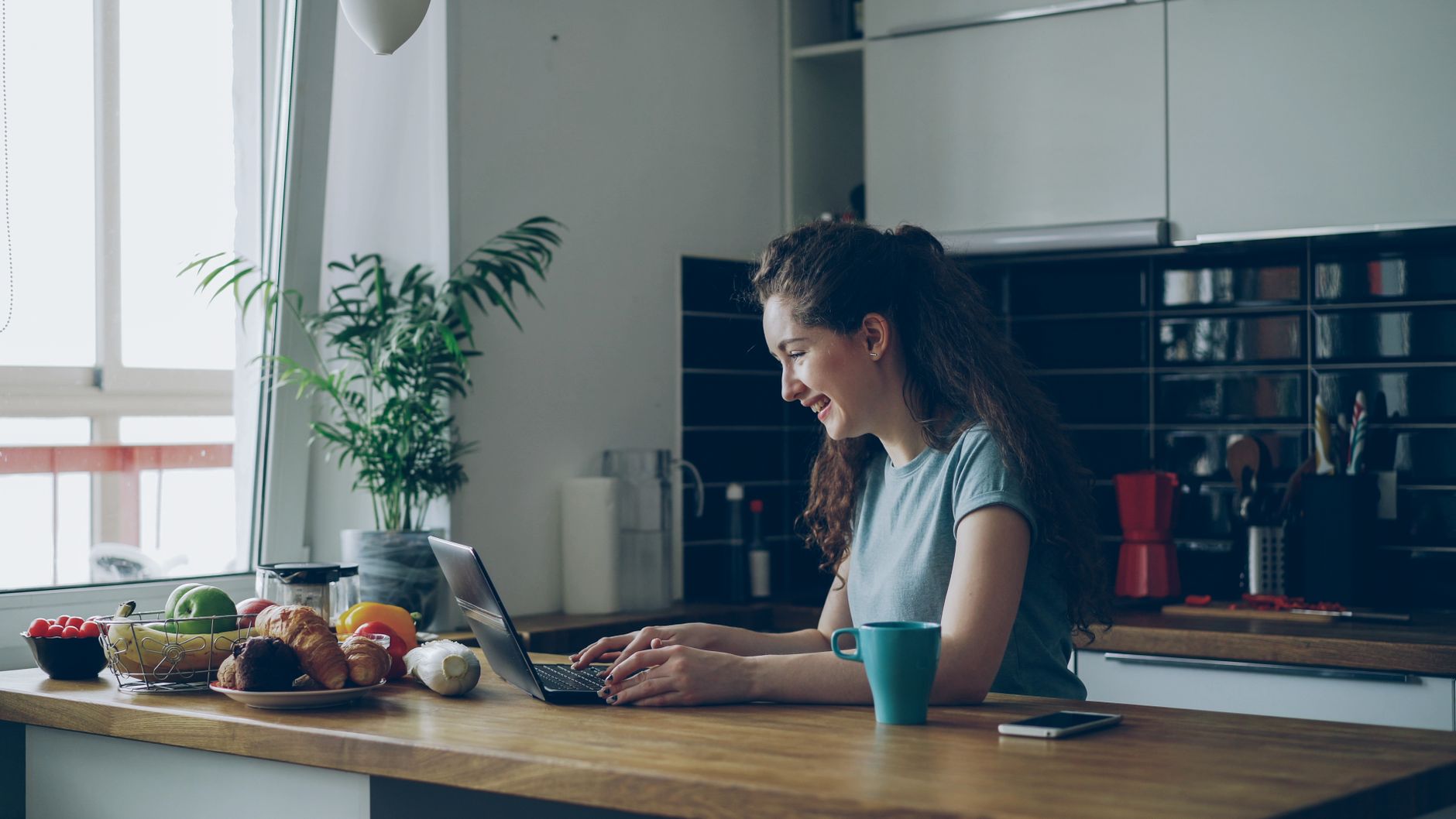 A woman working on a laptop at a kitchen island with a plant, a mug, and a bowl of fruit