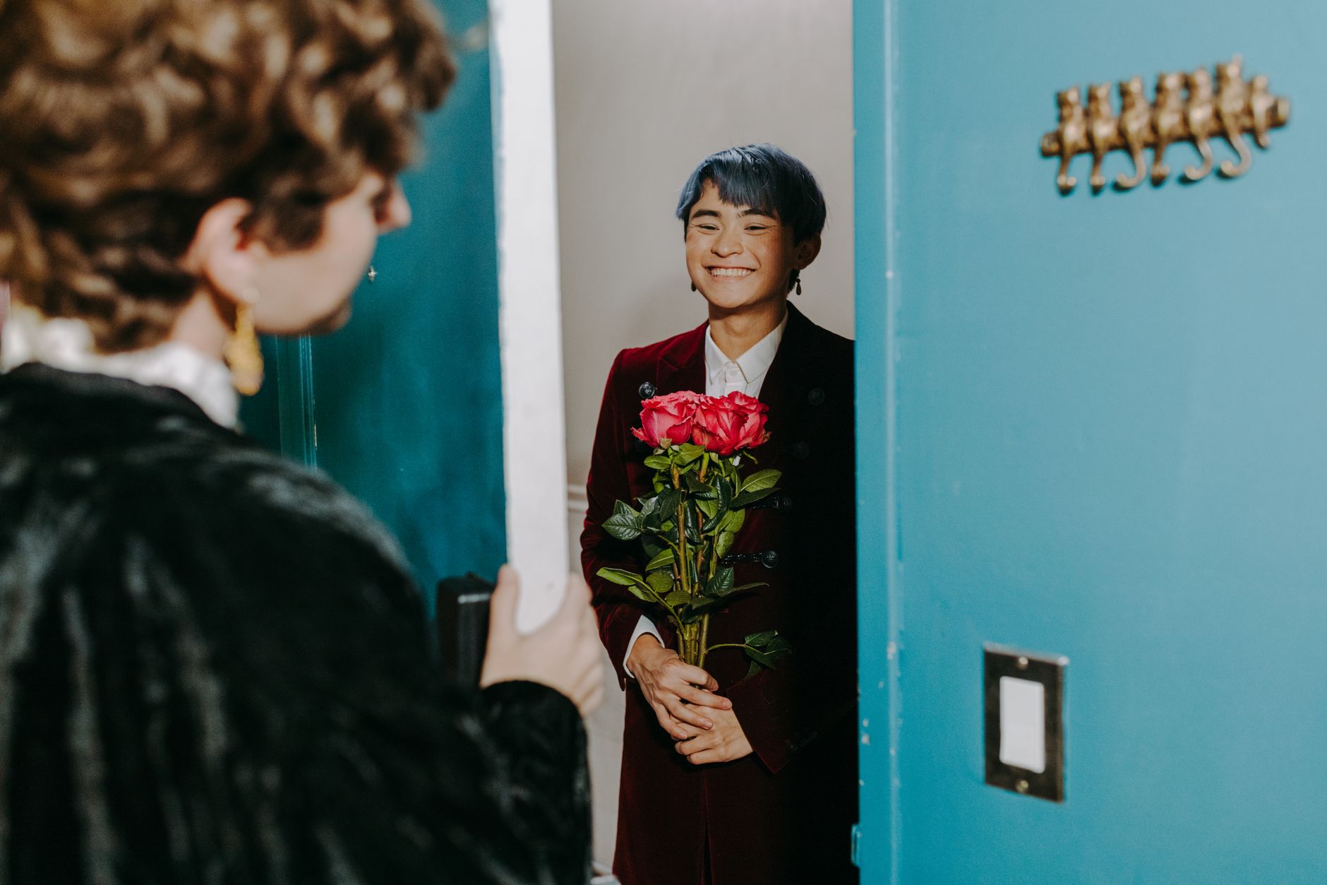 A smiling person in a burgundy blazer holding a bouquet of pink roses at a teal doorway