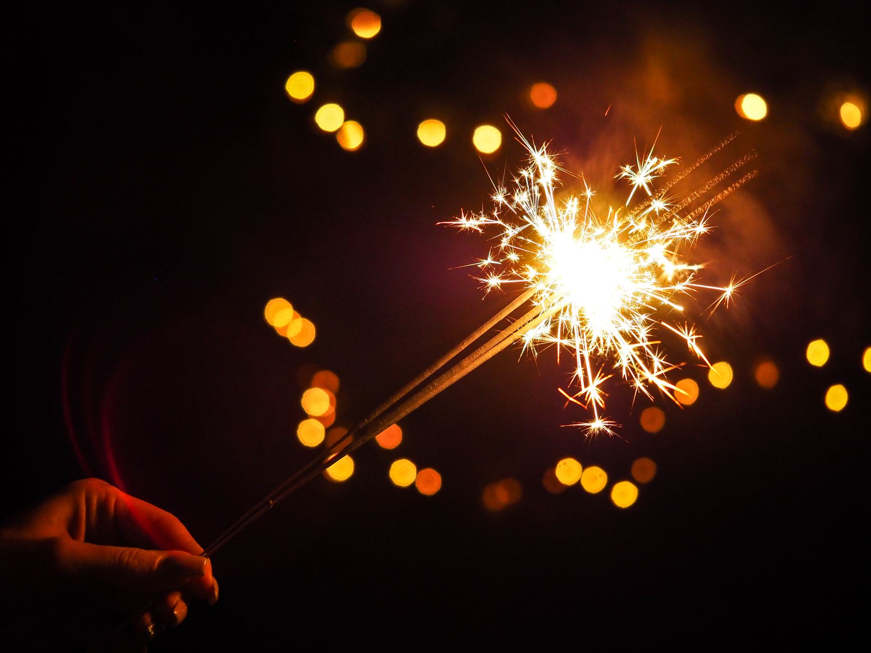 A hand holding a glowing sparkler against a dark background with golden bokeh lights