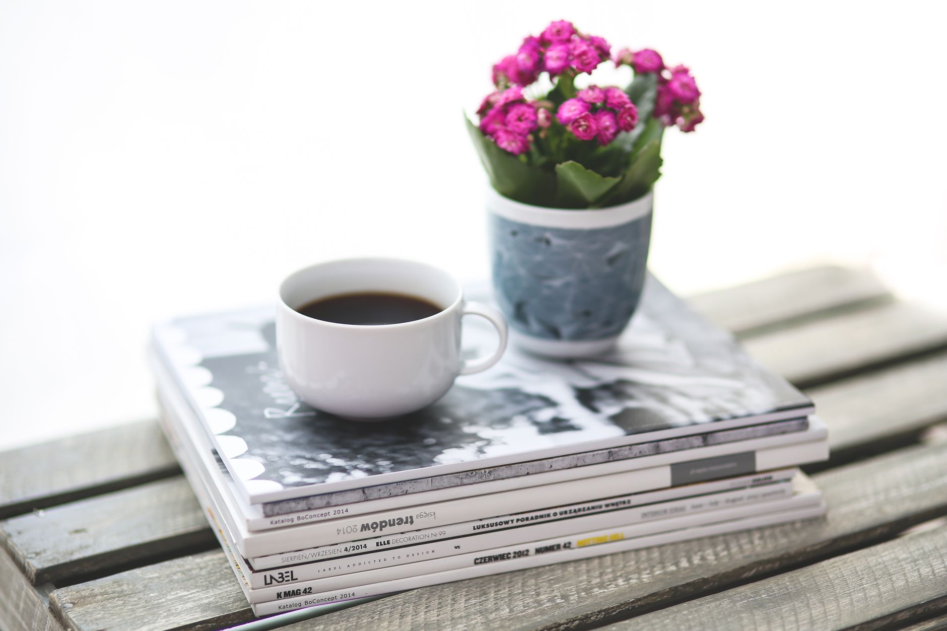 A cup of coffee on a stack of magazines beside a small pot of pink flowers