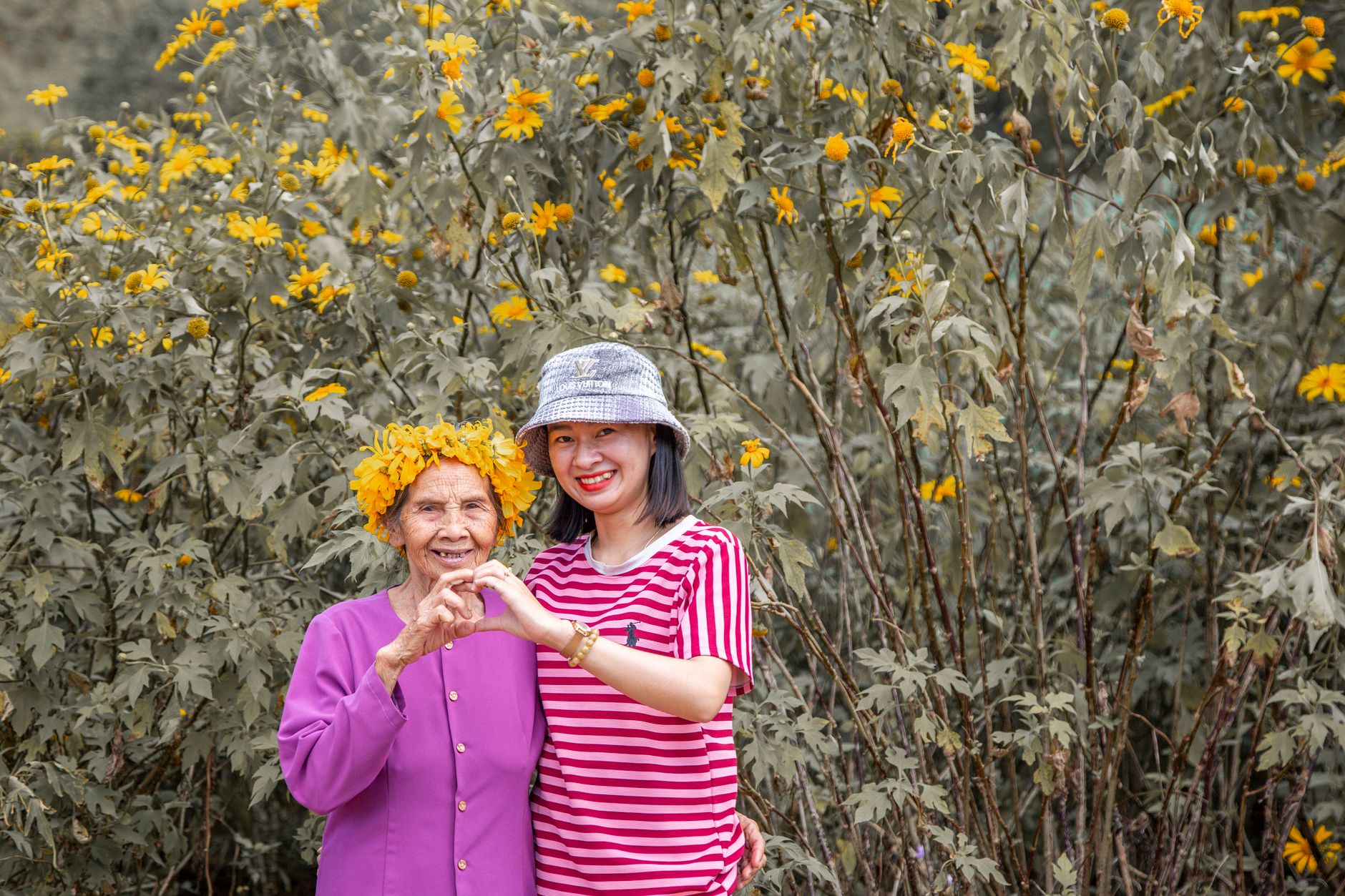 A grandmother wearing a yellow flower crown and her granddaughter making a heart with their hands in front of yellow flowers