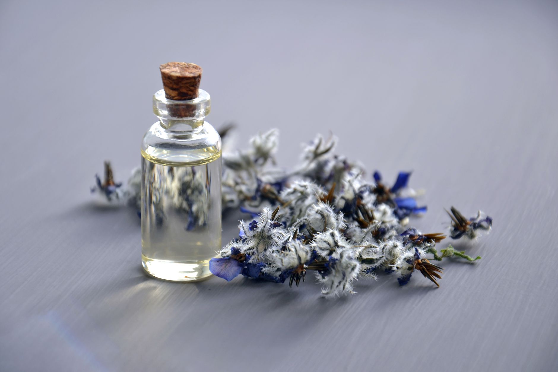 A small corked glass bottle of essential oil resting beside a bundle of dried purple lavender-toned flowers