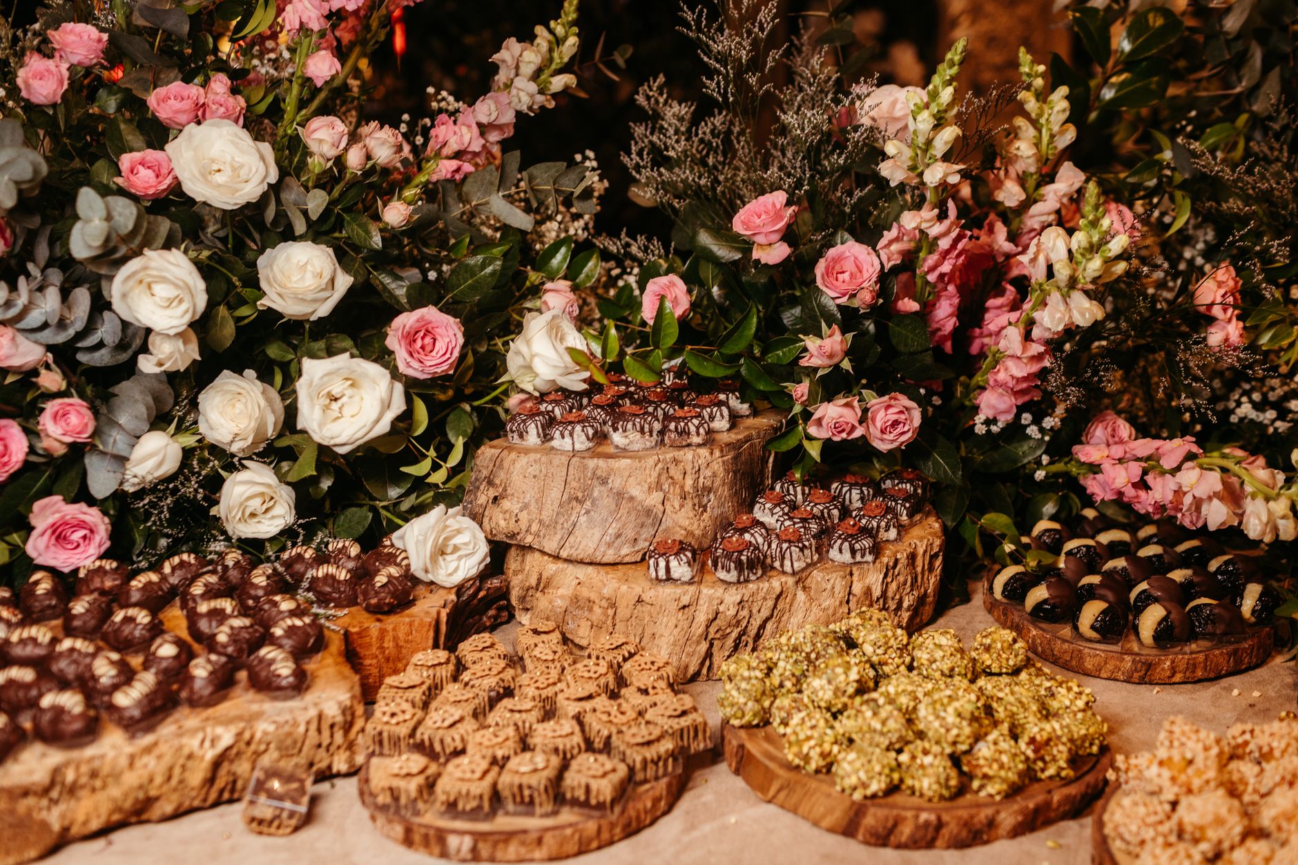 A dessert table of chocolates and sweets on wooden slabs surrounded by pink roses, white blooms, and greenery