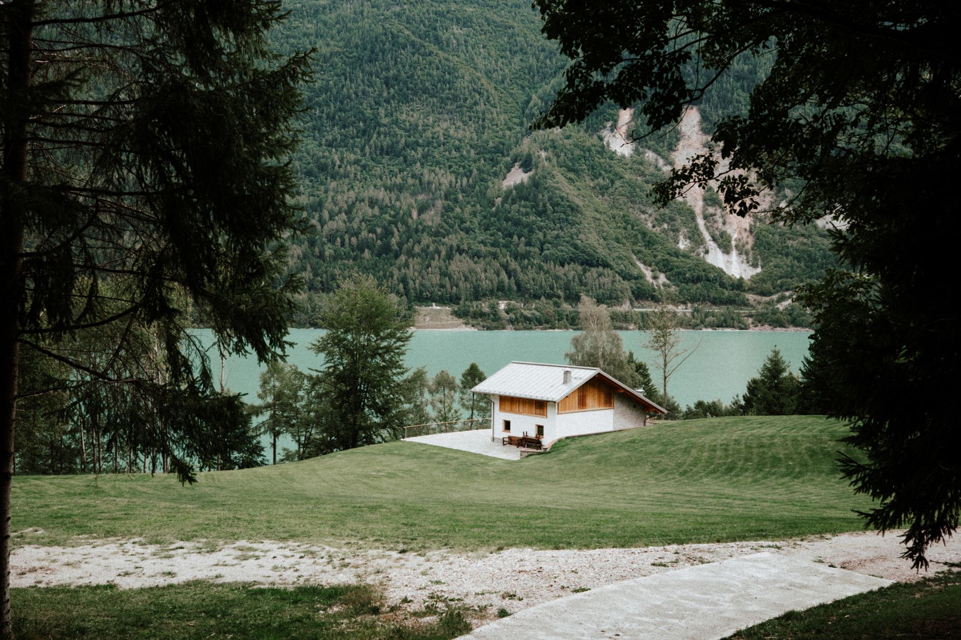 A small wooden cabin on a green lawn above a turquoise mountain lake surrounded by forest