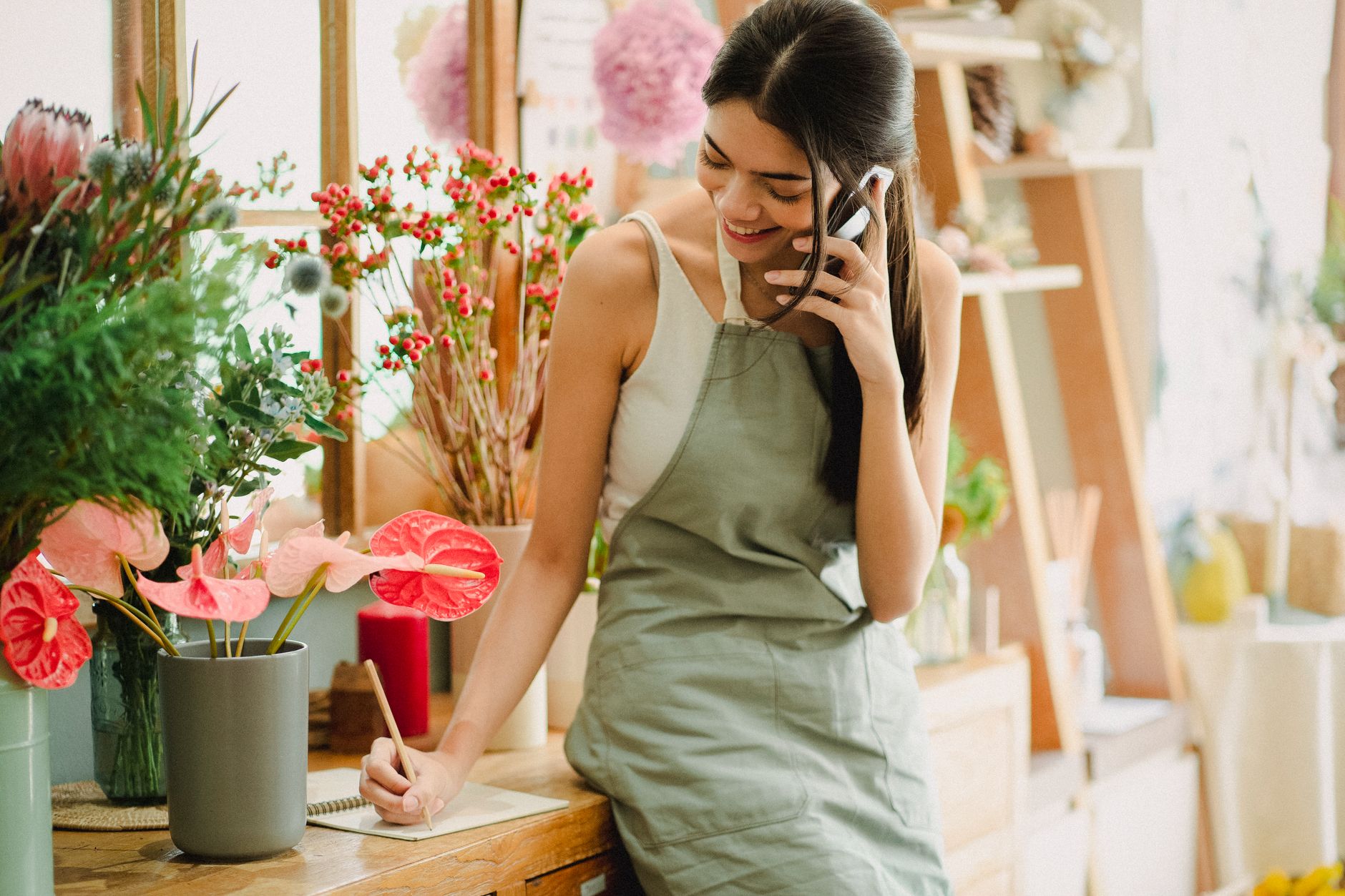 A smiling florist in a gray apron taking an order on her phone and writing notes at the counter of a flower shop
