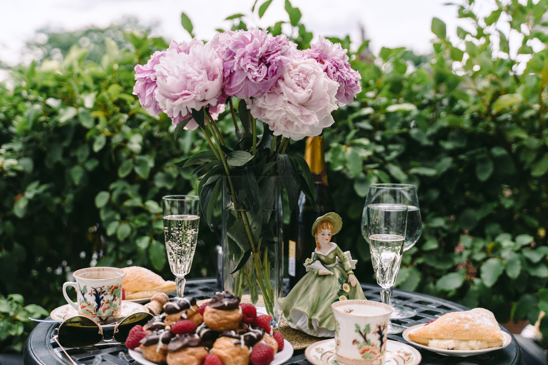 A vase of pink peonies on a garden table beside pastries, tea cups, and glasses of sparkling wine