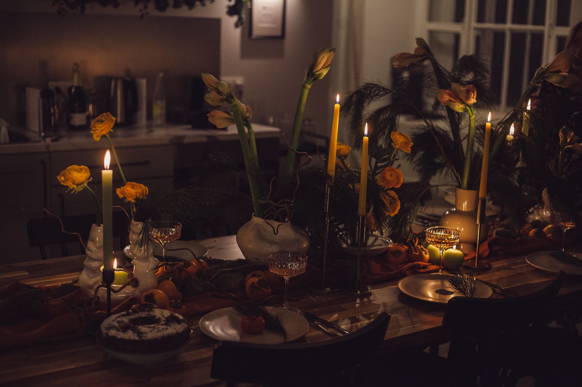 Una mesa de comedor a la luz de las velas con ran&uacute;nculos naranjas, tulipanes y calabazas peque&ntilde;as en un ambiente c&aacute;lido de oto&ntilde;o