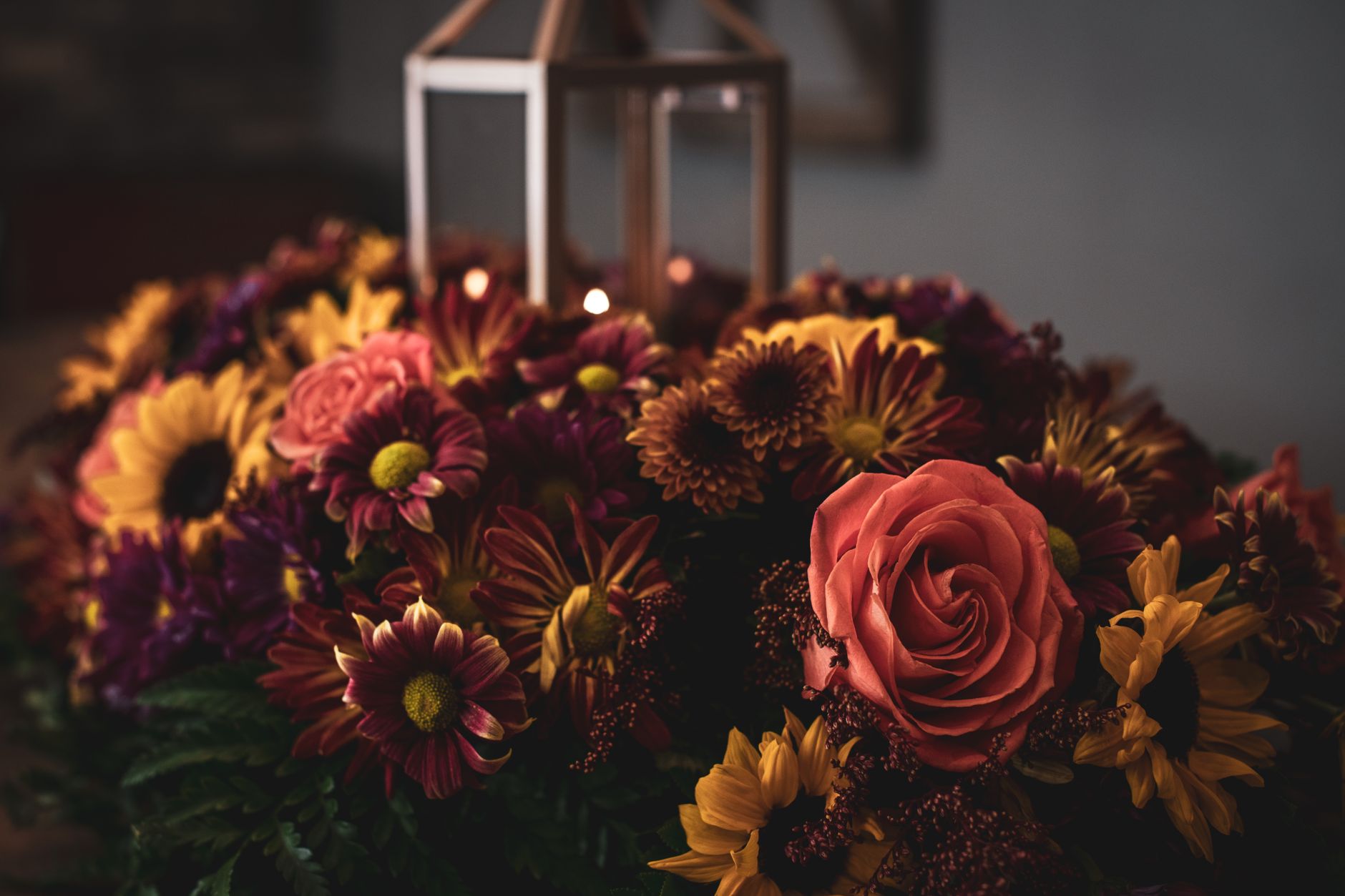 A dense autumn arrangement of burgundy and gold chrysanthemums with a coral rose and a lantern glowing behind it