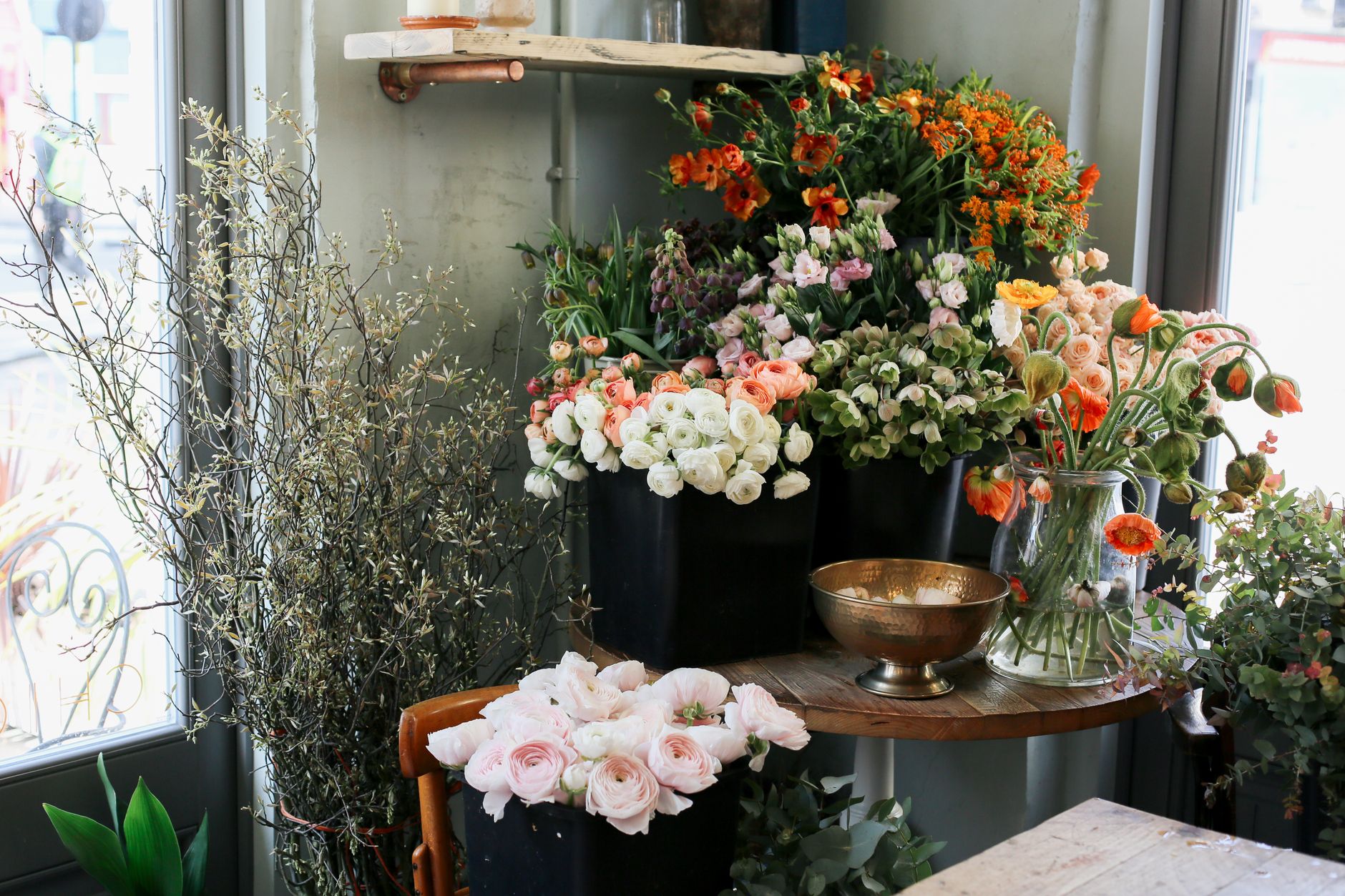A cozy flower shop window display with pink ranunculus, orange blooms, and white flowers in vases and pots