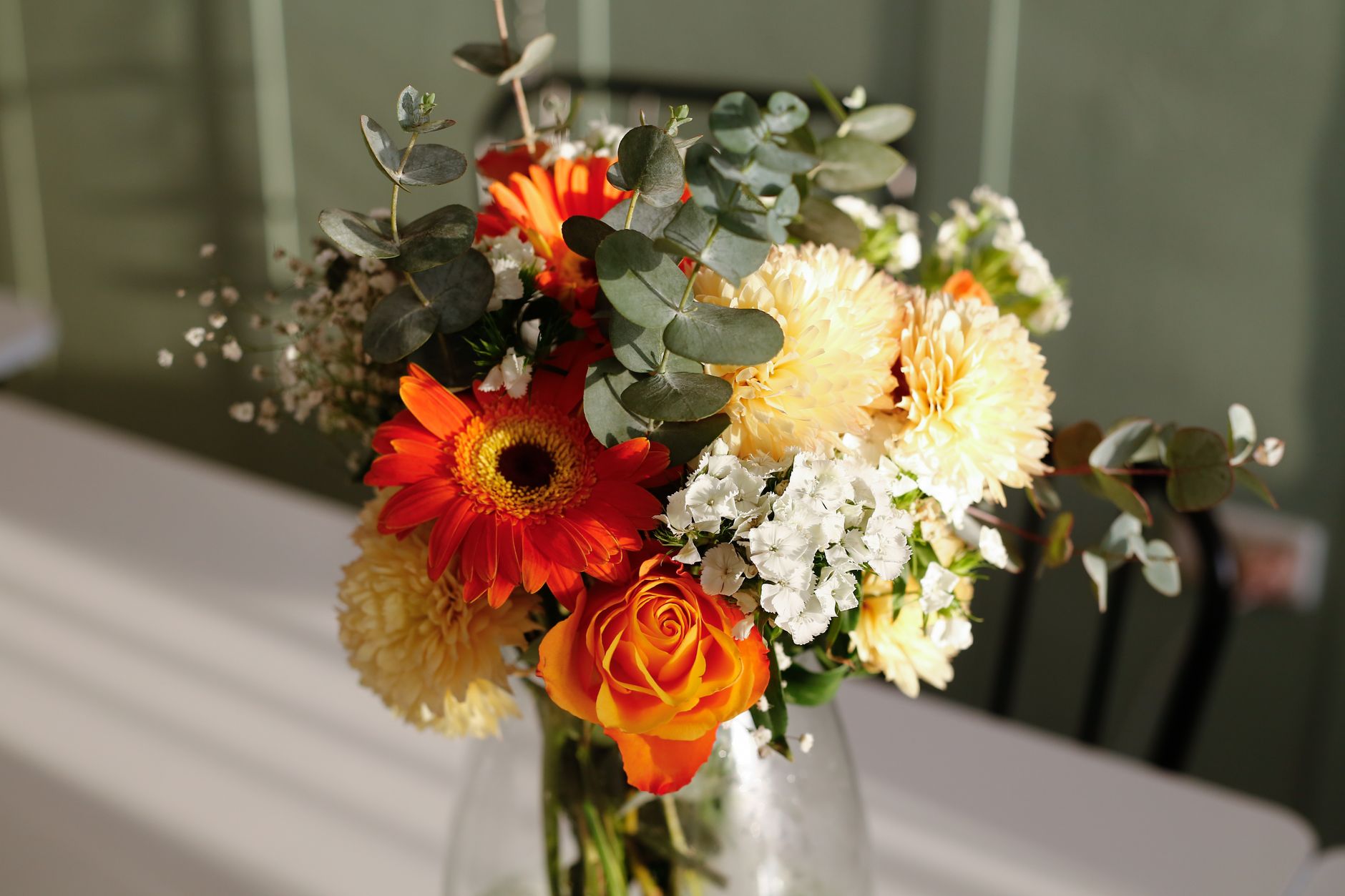 A mixed bouquet of orange gerbera, a rose, cream dahlias, white filler flowers, and eucalyptus in a glass vase on a sunlit table