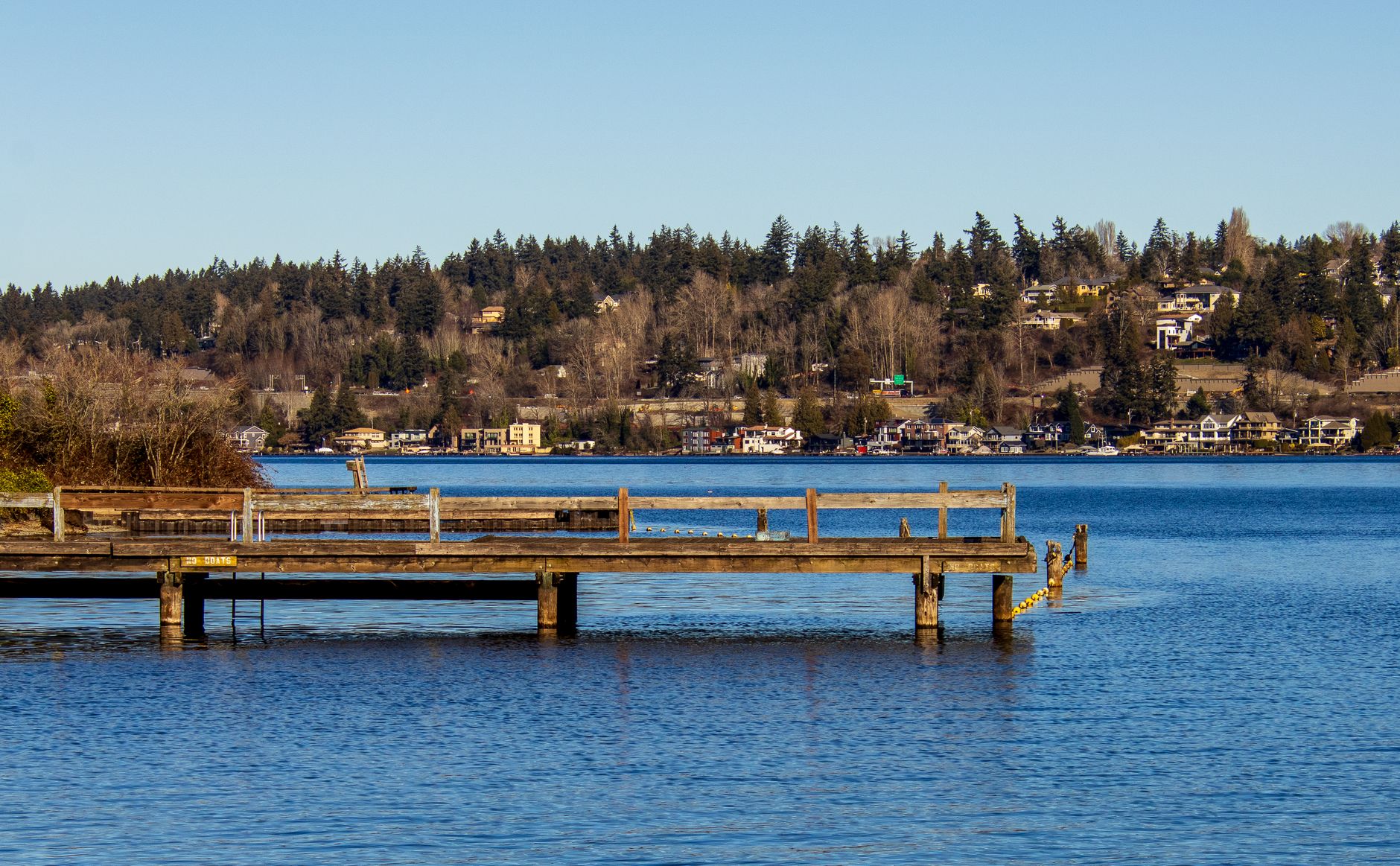 A wooden dock on a calm blue lake with homes set among evergreen trees on the wooded hillside beyond