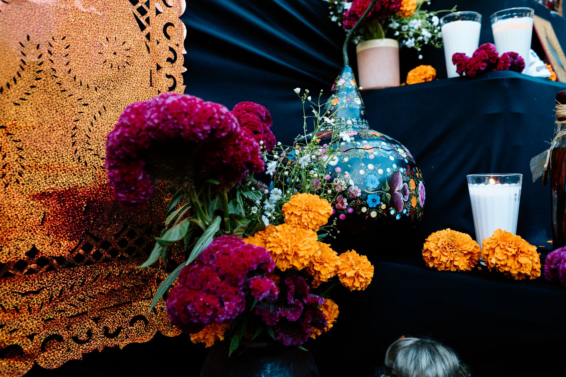 An ofrenda with orange marigolds, deep magenta cockscomb, small white flowers, white candles, and golden cut paper on a black draped altar / Una ofrenda con flores naranjas, una flor magenta de terciopelo, flores blancas, velas blancas y papel picado dorado sobre un altar con tela negra