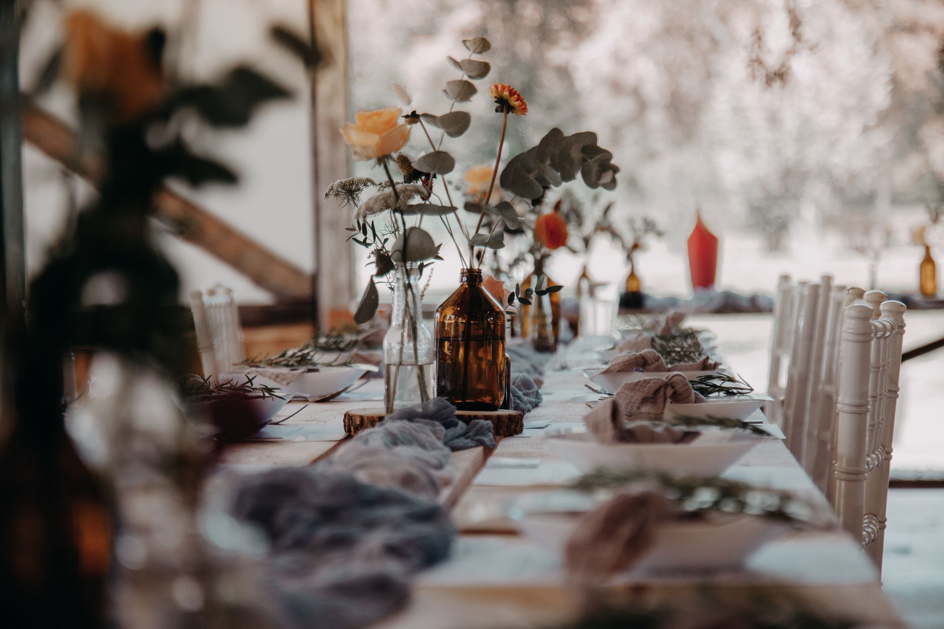 A long rustic dinner table set with several small glass vases of flowers and greenery down its length