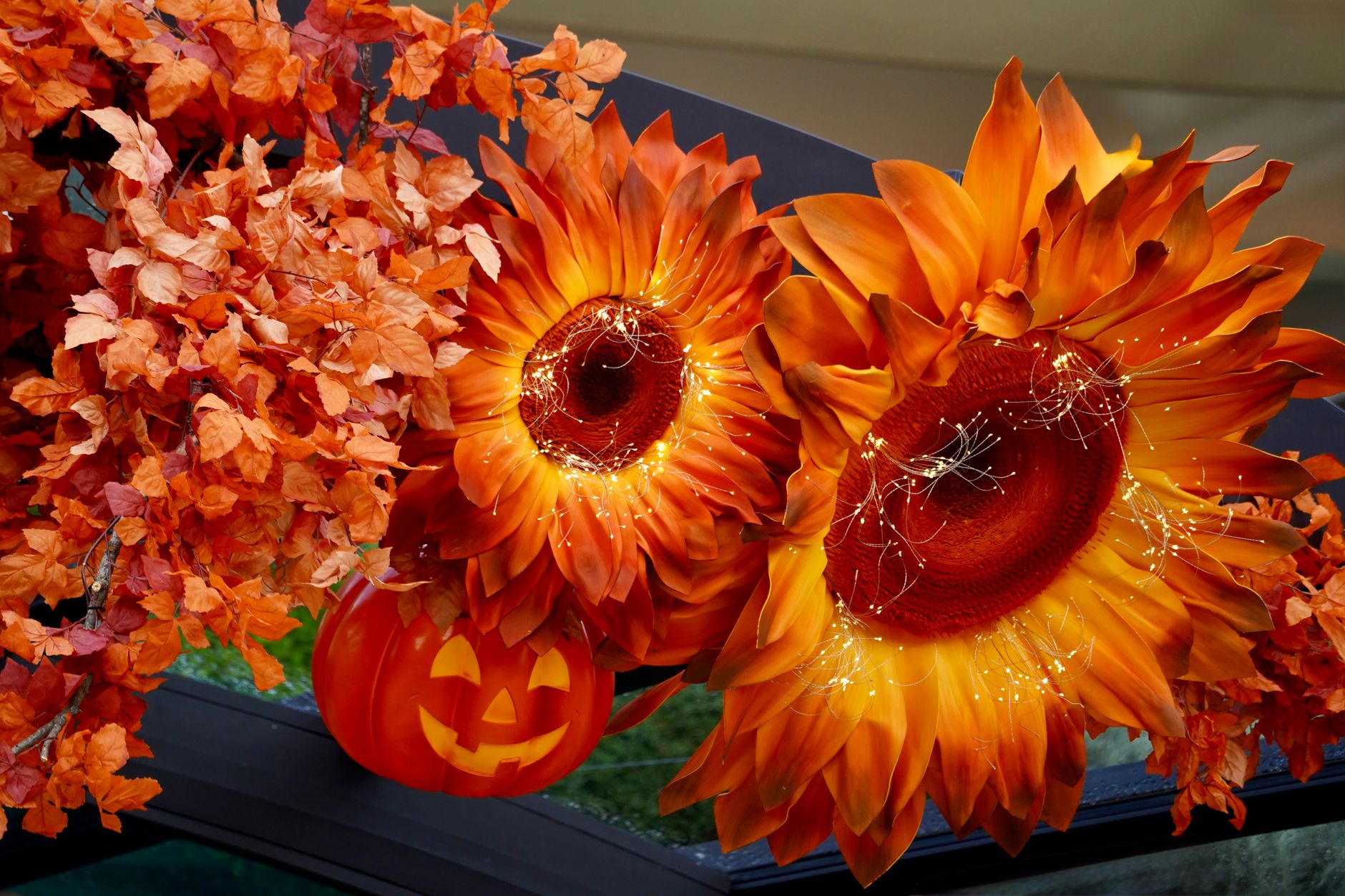 Orange sunflowers wrapped in tiny string lights beside a carved jack-o-lantern and orange autumn leaves